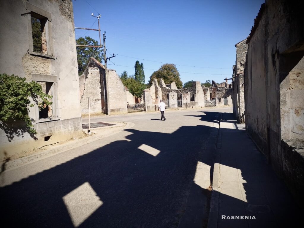photo taken at Oradour-sur-Glane. A man is crossin the road beneath a blue sky. The buildings are in ruins, though many walls still stand. A large black shadow looms in the foreground.