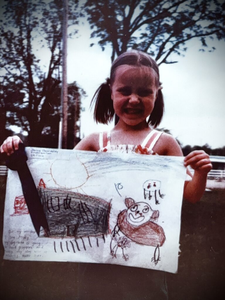 photo of the author circa 1978 standing outdoors and holding before her a child's drawing of a dog with a goofy grin and mouse ears next to a boxy black building beneath a sunny blue sky.