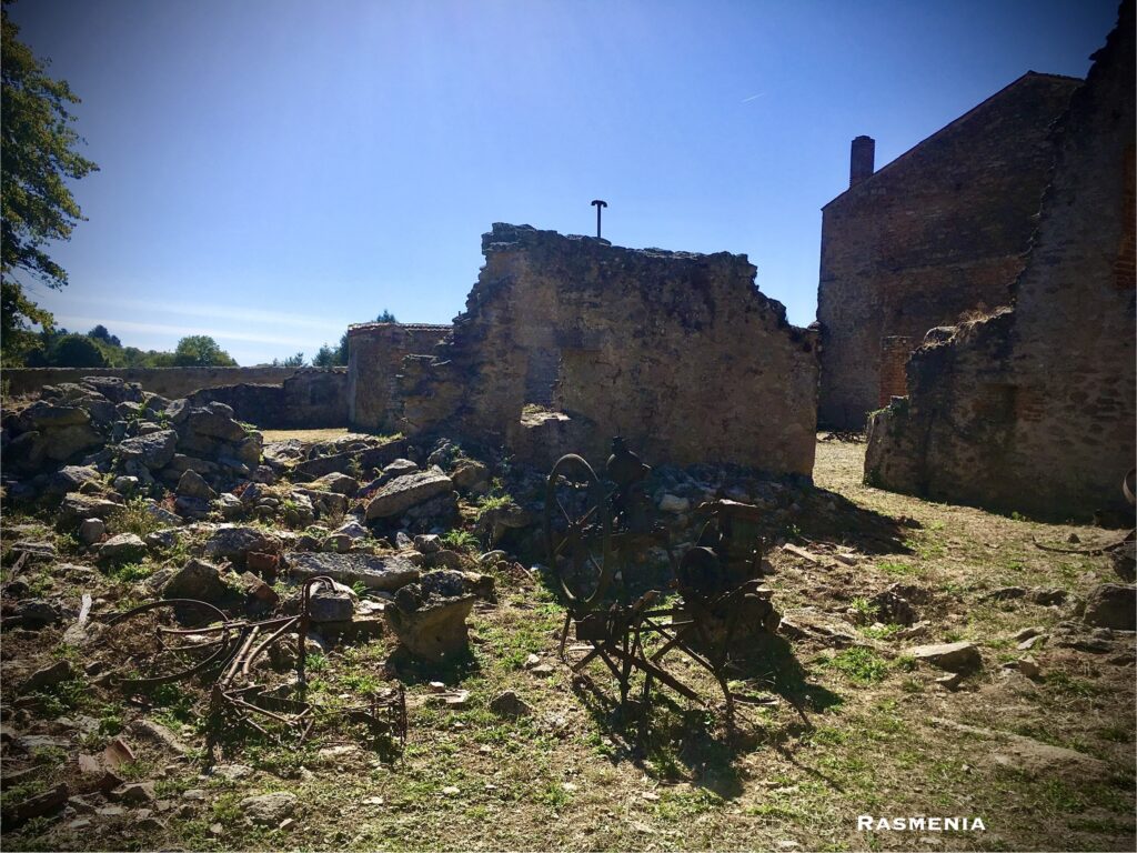 the ruins at Oradour-sur-Glane. The sky is blue above, and several scattered rusty objects of daily life such as a bicycle and a spinning wheel rest in the grass and rocks below.