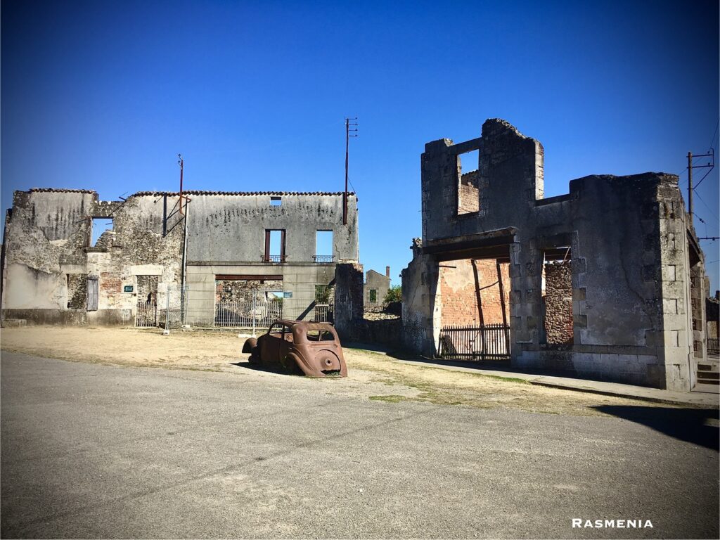 the skeleton of an old rusted car rests in the street of Oradour-sur-Glane, the hollowed out buildings long burned stand behind it beneath a vibrant blue sky.